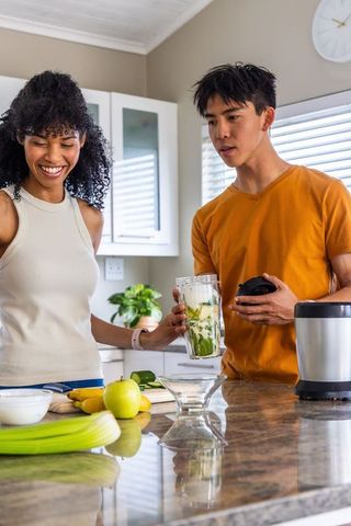 Diverse couple making green smoothie on kitchen island with blender and fresh produce