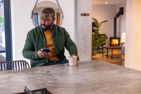 African american man checking phone while holding coffee at modern cozy kitchen countertop interior