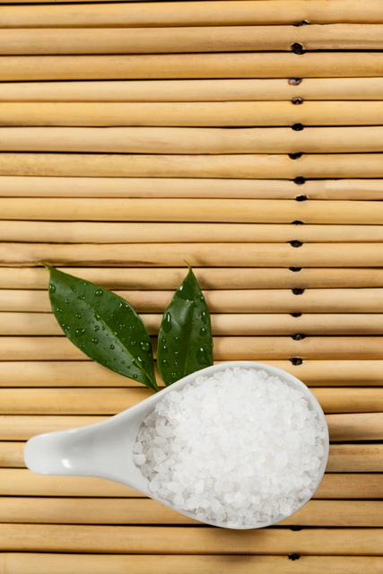 Sea salt in a white spoon with green leaves on a bamboo mat, perfect for illustrating concepts of natural wellness, spa treatments, and relaxation. Ideal for use in health and beauty blogs, eco-friendly product advertisements, and holistic therapy promotions.
