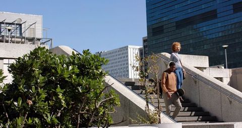 Young friends walking down concrete steps holding skateboard by glass tower in daylight