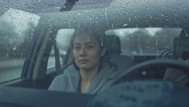 Asian woman sitting in car looking through rain-speckled windshield on overcast day