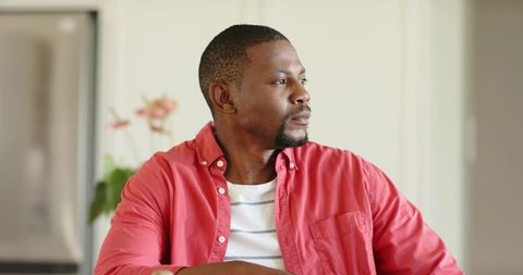 African american man gazing thoughtfully while sitting in kitchen wearing red shirt