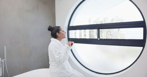Woman in bathrobe enjoying warm beverage by large modern window