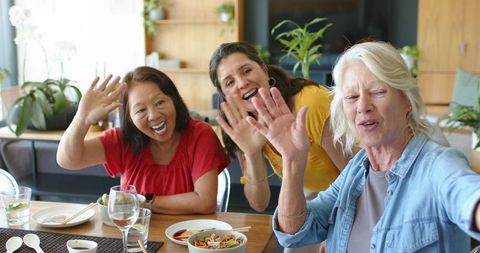Diverse friends waving and taking selfie around cozy dining table during casual meal