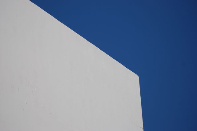 This image depicts a minimalist white wall set against a clear blue sky. The sharp geometric lines and the stark contrast between the white wall and the blue sky embody simplicity and modern architectural aesthetics. This image is ideal for use in design projects that need a clean, uncluttered background or for conveying themes of modern living, minimalism, and elegance.