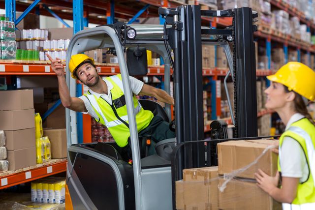 Male worker operating forklift in warehouse, moving boxes with another worker guiding. Suitable for topics on logistics, teamwork, industrial safety, supply chain management, and warehouse operations.