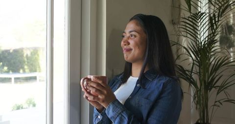 Serene woman holding mug gazing out sunlit window in cozy home interior
