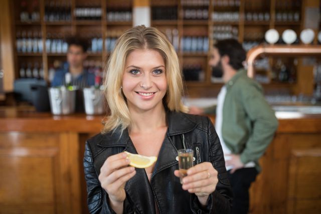 Blonde woman smiling while holding a tequila shot and lemon slice in a bar. Ideal for use in advertisements for nightlife, social events, bars, and alcoholic beverages. Perfect for illustrating themes of fun, celebration, and socializing.