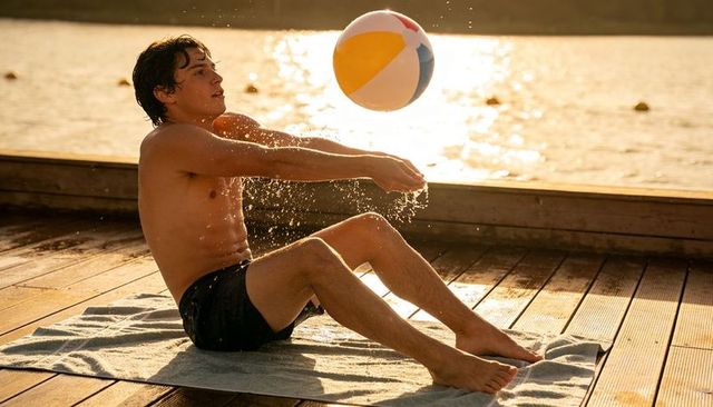 Shirtless young man playing with colorful beach ball on wooden lakeside dock at golden hour