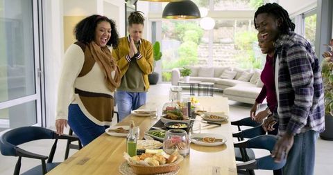 Multiracial friends laughing and gathering around long wooden dining table, sharing homemade meal