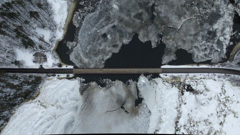 Aerial bridge crossing frozen river with breaking ice and snow-covered forest landscape