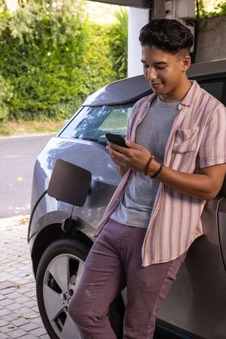 Young Man Browsing Smartphone Beside Charging Electric Car on Driveway
