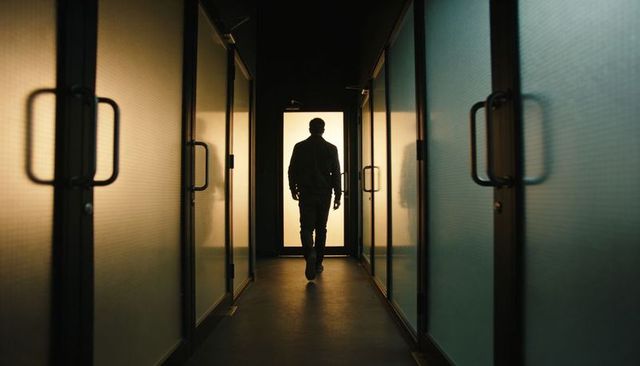 Lone man walking down narrow backlit office corridor with frosted glass partitions