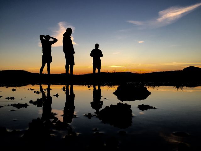 Silhouettes of three people standing near water with reflections capturing a stunning sunset. Use for travel, adventure, and nature themes. Perfect for promoting calm and serene experiences or travel destinations. Ideal for blogs, web banners, and social media posts about relaxation, outdoor activities, and scenic beauty.
