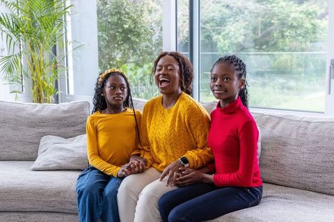 African american mother laughing with daughters holding hands on cozy sofa by sunny window