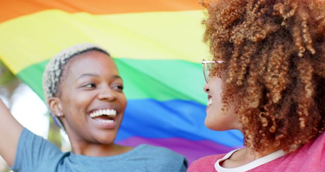 Happy diverse lesbian couple embracing and holding a rainbow flag outdoors. Togetherness, relationship, love, lgbtg awareness summer and nature, unaltered.