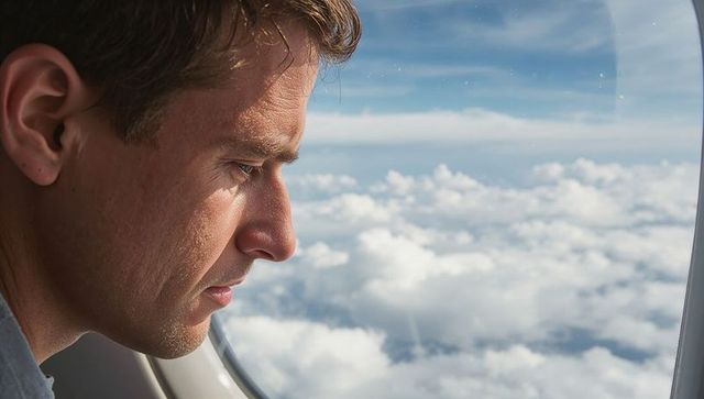 Adult man gazing contemplatively through plane window over cloudscape during daylight flight
