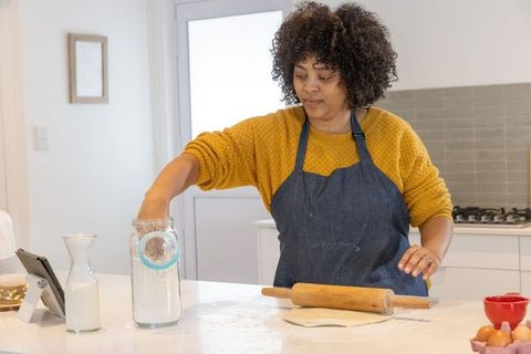 Home baker reaching into flour jar while rolling out dough with wooden rolling pin