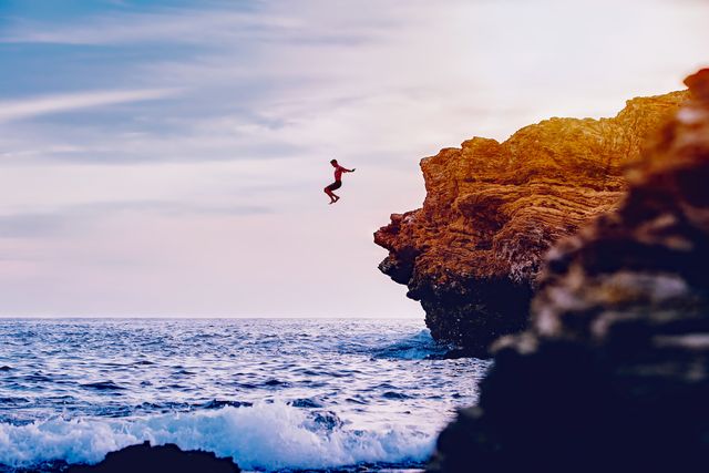Individual leaps off cliff into ocean with waves crashing below at sunset. Perfect for adventure concepts, travel promotions, extreme sports content, and thrill-seeking lifestyle features.
