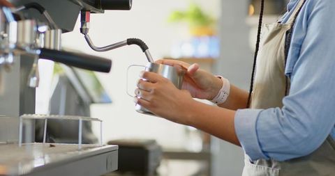 Barista frothing milk with steam wand at espresso machine close-up, hands holding pitcher