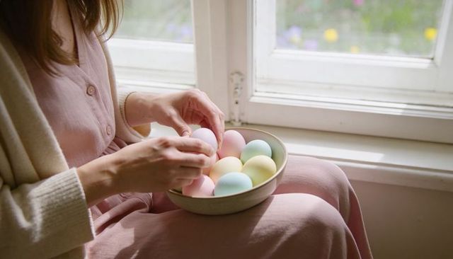 Hands holding ceramic bowl of pastel eggs on sunlit lap by window, cozy minimalist spring