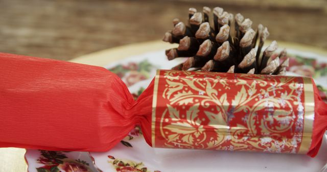 Traditional holiday cracker in vibrant red and gold sitting on an ornate floral plate with a pinecone in the background; perfect for holiday event invitations, seasonal promotional materials, or decorating festive party announcements.