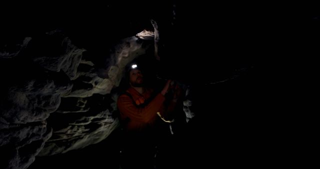 Caucasian man wearing headlamp exploring dark natural cave system looking up at rock, copy space. Exploration, adventure, caving, nature, hobbies and outdoor activities, unaltered.