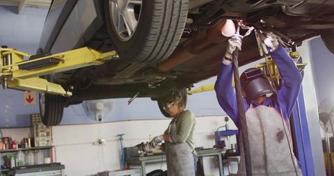Female mechanic welding beneath raised car on auto lift in busy garage workshop
