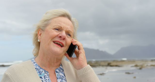 Elderly woman enjoying a peaceful moment by the sea, speaking on the phone during an overcast day. Perfect for concepts related to retirement, relaxation by nature, communication, and senior lifestyle. Ideal for use in health and wellness articles, senior living promotions, and telecommunication advertisements.