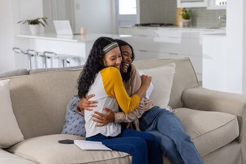 Sisters hugging on beige sofa celebrating joyful news with letter, smiling in modern home