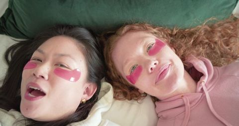 Two women lying on bed wearing pink under-eye gel patches and cozy pastel hoodies