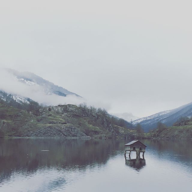 Misty morning with a solitary cabin on a calm mountain lake surrounded by fog-draped hills. Ideal for promoting relaxation, nature retreats, or meditation content. Suitable for travel brochures and environmental awareness campaigns emphasizing tranquility and remoteness.