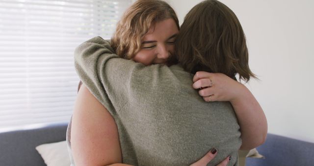 Two women hugging in a cozy living room with natural light. The scene expresses love, comfort, and emotional support, perfect for illustrating friendship, empathy, and human connection. Ideal for use in articles or advertisements focused on mental health, relationships, and community support.