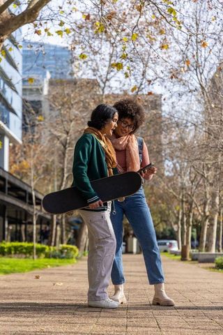 Two young women checking smartphone while holding skateboard on urban autumn campus
