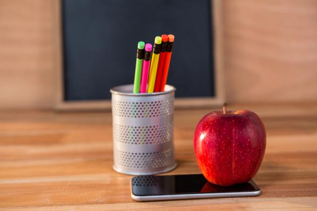 Red apple placed on a mobile phone next to a pencil holder with colorful pencils on a wooden desk. Blackboard in the background. Ideal for educational content, back-to-school promotions, classroom decor, and learning materials.