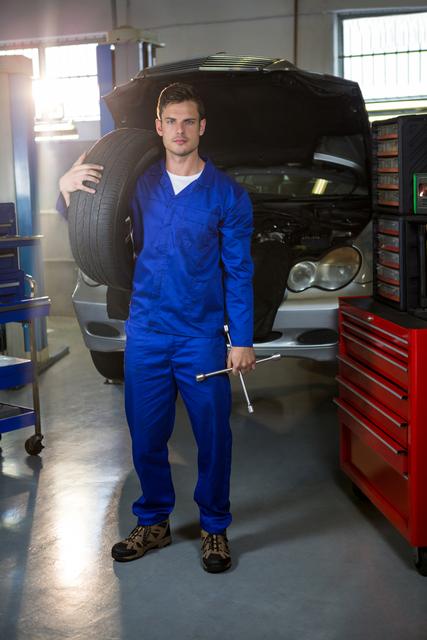 Portrait of mechanic holding tyre and lung wrench at repair garage