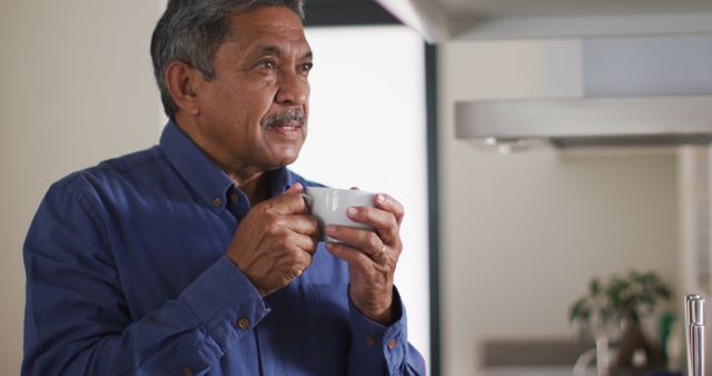 Mature man drinking coffee in kitchen, looking thoughtful and relaxed. Ideal for themes about seniors, everyday life, relaxation, thoughtful moments, cozy mornings, and domestic settings. Useful in lifestyle blogs, retirement articles, wellness promotions, and coffee brand advertisements.