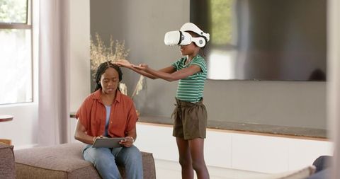 African American mother using tablet while daughter wearing VR headset in sunlit living room