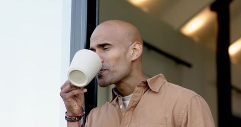 Mature bald man sipping coffee at doorway wearing casual button-up and beaded bracelet