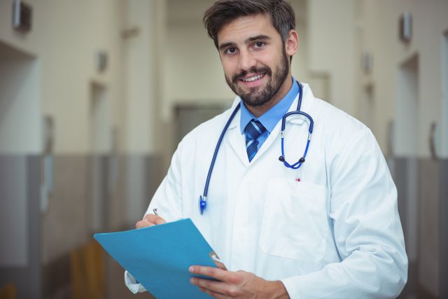 This image shows a male doctor in a hospital corridor, writing on a clipboard. He is smiling and wearing a white coat with a stethoscope around his neck. This image can be used for healthcare, medical, and professional themes, including hospital websites, medical brochures, and healthcare advertisements.