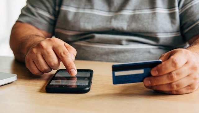 Middle-aged man making secure mobile payment with credit card and smartphone at home desk