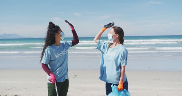 Two diverse women wearing volunteer t shirts and face masks picking up rubbish from beach. eco conservation volunteers, beach clean-up during coronavirus covid 19 pandemic.