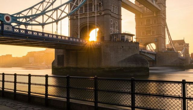Sunburst through tower bridge at golden hour with riverside reflections and promenade rail