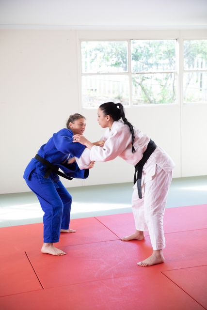 Two Caucasian female judokas wearing blue and white judogi, practicing judo on a mat during a training in a bright studio.