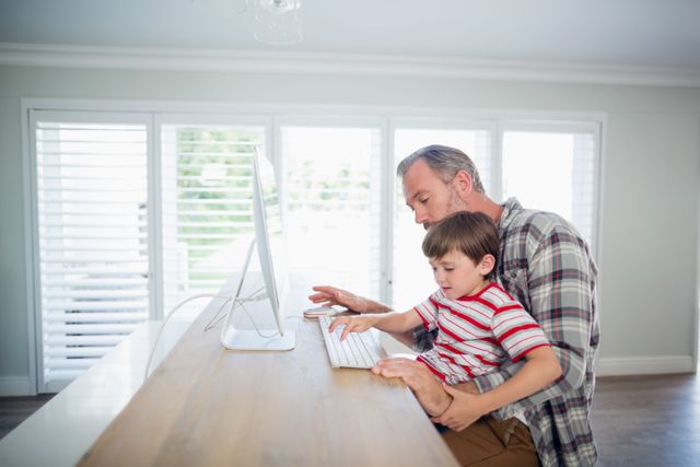 Father and son sitting at desk using computer together. Ideal for concepts of family bonding, modern parenting, home education, and work-life balance. Suitable for articles, blogs, and advertisements related to family life, technology in the home, and educational activities.