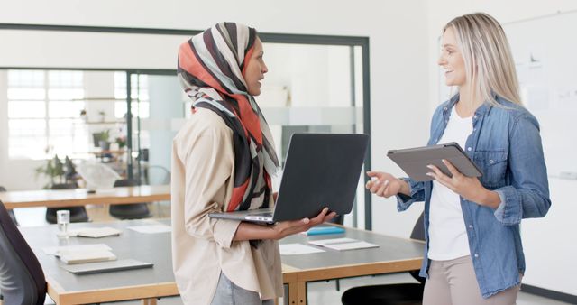 Two professional businesswomen are standing and discussing work in a modern office. One woman is holding a laptop while the other is holding a tablet. They are smiling and appear to be engaged in a productive conversation. Suitable for use in contexts involving workplace diversity, teamwork, professional collaboration, and modern office environments.