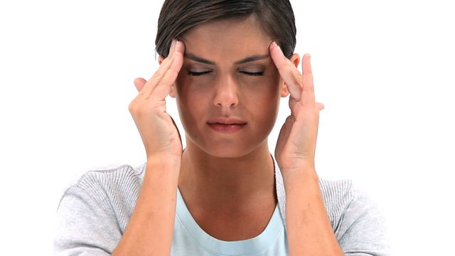 The scene captures a young brunette woman placing her hands on her temples, indicating she might be experiencing a headache or stress. The expression is one of discomfort or concentration. The simplicity of the white background reinforces a focus on health-related themes. Ideal for articles and websites about headache relief, stress management, or mental health awareness.