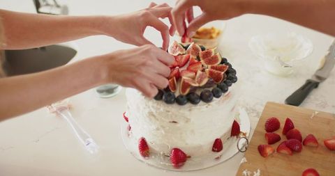 Two friends decorating layered homemade cake with figs strawberries and blueberries