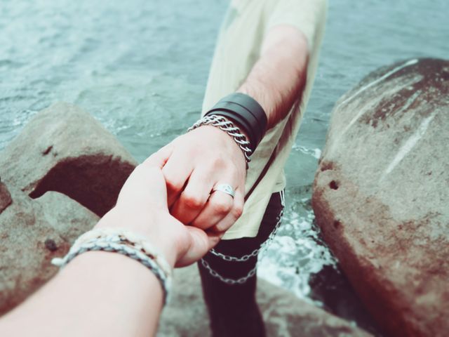 Two individuals are holding hands near ocean rocks, symbolizing commitment and love. One person is wearing chain bracelets, indicating a stylish and modern look. Ideal for themes around love, relationship, romance, togetherness, and travel stories.