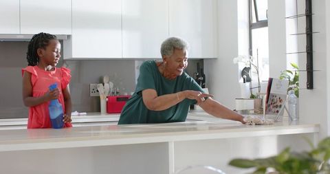 African-american grandmother and granddaughter cleaning bright modern kitchen, wiping counter and bo